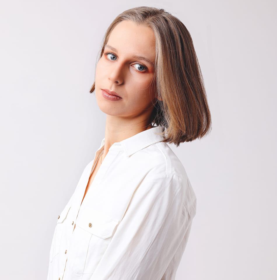 Na zdjęciu widać osobę w białej koszuli, jest ustawiona bokiem do kamery. Ma niebieskie oczy, jasnobrązowe włosy do ramion. Lekko się uśmiecha.